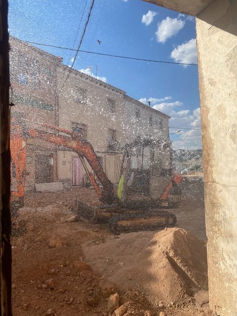 View through window of construction site with red machinery and dirt on ground, mountains visible in distance under blue sky