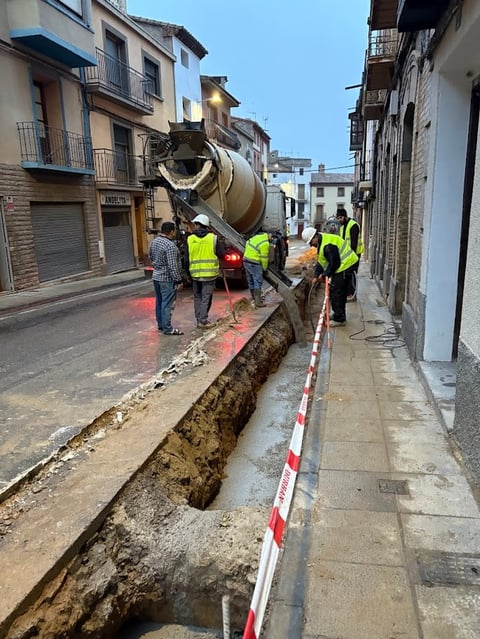 Construction workers in safety vests operate a cement mixer truck on a narrow European street with exposed underground pipes and red/white warning tape.